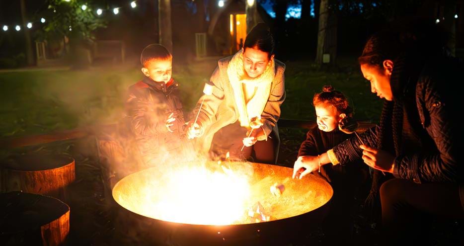 A family toasting marshmallows by the firepit at Pinewood glamping pods at Port Lympne Hotel & Reserve in Kent, UK